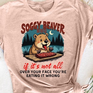 Könnte beinhalten: Ein hellbraunes T-Shirt mit einem Cartoon-Biber, der ein Steak isst. Der Text "Soggy Beaver BBQ" befindet sich über dem Biber und der Text "if it's not all over your face you're eating it wrong" befindet sich unter dem Biber.