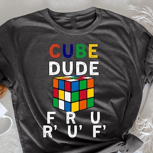 Może przedstawiać: Koszulka w kolorze ciemnoszarym melanż z napisem "CUBE DUDE" i grafiką kostki Rubika. Pod kostką znajduje się napis "F R U R' U' F'.