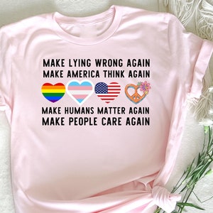May include: Pink t-shirt with the text "MAKE LYING WRONG AGAIN, MAKE AMERICA THINK AGAIN, MAKE HUMANS MATTER AGAIN, MAKE PEOPLE CARE AGAIN". The text is in black and there are four hearts with different designs: a rainbow heart, a transgender heart, an American flag heart, and a peace sign heart.
