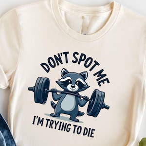 以下が含まれることがあります： クリーム色のTシャツに、バーベルを持ち上げる漫画のラクーンが描かれています。Tシャツの文字は「Don't Spot Me I'm Trying To Die.」です。