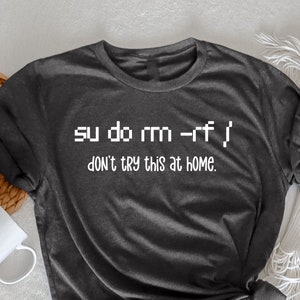 May include: Dark grey heather t-shirt with the text "su do rm -rf / DON'T TRY THIS AT HOME." A white mug and a woven basket are in the background. The shirt is folded and tied at the bottom.