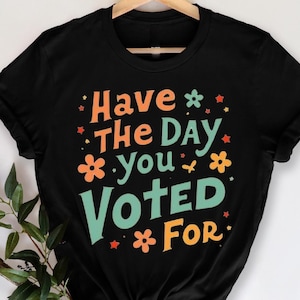 Op de afbeelding: Zwart T-shirt met de tekst "Have The Day You Voted For" in oranje, groen en geel. Het ontwerp bevat bloemen- en sterrenafbeeldingen. Het shirt hangt aan een houten hanger.