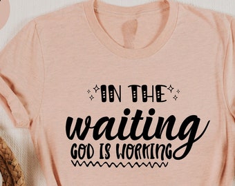 May include: Heather peach-colored t-shirt with the text "IN THE waiting GOD IS WORKING" in black script. The shirt is folded, with a knot at the bottom. A cup of coffee, a small plant, and a notebook are visible.