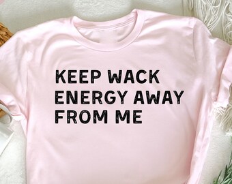 May include: A light pink t-shirt with the text "KEEP WACK ENERGY AWAY FROM ME" in black. The shirt is knotted at the bottom and is displayed on a white surface with a sprig of white flowers.