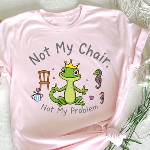 Pode incluir: Camiseta rosa com um gecko verde usando uma coroa e o texto "Not My Chair. Not My Problem."