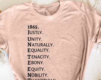 May include: Heather peach-colored t-shirt with the text "1865. JUSTLY. UNITY. NATURALLY. EQUALITY. TENACITY. EBONY. EQUITY. NOBILITY. THANKFULLY. HONORARY." The shirt is folded and tied at the bottom. A white mug and flowers are in the background.
