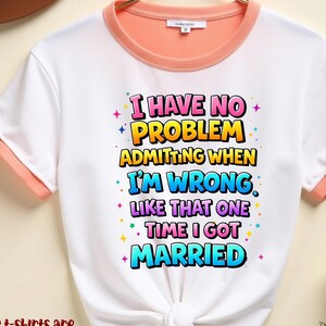 May include: White t-shirt with peach-colored trim and the text "I HAVE NO PROBLEM ADMITTING WHEN I'M WRONG LIKE THAT ONE TIME I GOT MARRIED" in colorful bubble letters. The shirt is hanging on a gold hanger.