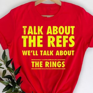 May include: Red t-shirt with yellow text that reads "Talk About The Refs We'll Talk About The Rings"