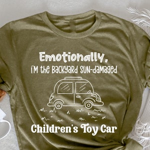 Könnte beinhalten: Heather olivgrünes T-Shirt mit weißem Text und einer Autografik. Der Text lautet "Emotionally, I'M the Backyard SUN-damaged Children's Toy Car". Das Shirt ist auf einer weißen Oberfläche gefaltet.