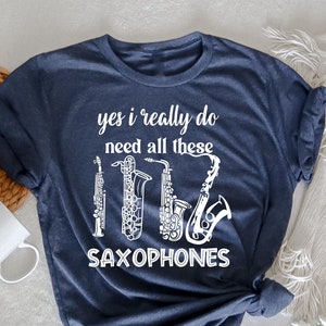 Op de afbeelding: Een marineblauw t-shirt met de tekst "yes i really do need all these saxophones" in wit. De tekst wordt omringd door een afbeelding van vier saxofoons.