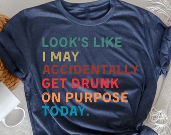 May include: Heather navy blue t-shirt with the text "LOOK'S LIKE I MAY ACCIDENTALLY GET DRUNK ON PURPOSE TODAY." The text is in various colors. A white mug and a woven basket are in the background.