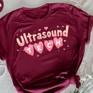 Op de afbeelding: Bordeauxrood T-shirt met de tekst "Ultrasound TECH" in een speels lettertype, met het woord "TECH" in roze hartvormen. Het shirt is aan de zoom geknoopt. Een witte mok en bloemen staan op de achtergrond.