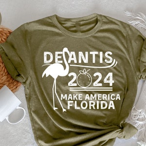May include: Heather olive green t-shirt with white text and a flamingo graphic. The text reads "DeSantis 2024 Make America Florida". A white coffee mug and woven basket are in the background.