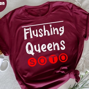 May include: Maroon t-shirt with the text "Flushing Queens SOTO" in white and red. The shirt is folded and tied at the bottom. A white mug and flowers are in the background.