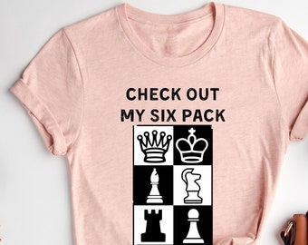 Può includere: T-shirt color pesca con la scritta "CHECK OUT MY SIX PACK" e una grafica di pezzi degli scacchi. I pezzi degli scacchi sono disposti in una griglia 2x3 di quadrati bianchi e neri. La maglietta è esposta su una superficie bianca.
