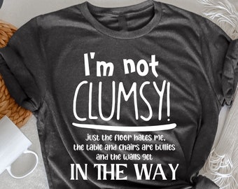 Puede incluir: Camiseta gris oscuro jaspeado con el texto "I'm not CLUMSY!" y la frase "Just the floor hates me, the table and chairs are bullies and the walls get IN THE WAY" en blanco. Una taza blanca y elementos decorativos en el fondo.