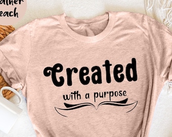 Puede incluir: Una camiseta color melocotón con las palabras "Created with a purpose" en escritura negra. El texto está encima de un diseño estilizado de un libro abierto. La camiseta está doblada y atada en la parte inferior, con una taza blanca y flores en el fondo.