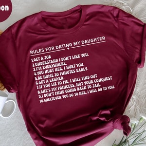 May include: Maroon t-shirt with white text listing rules for dating a daughter. The text includes phrases like "Get a job," "I'm everywhere," and "Get a lawyer." The shirt is folded and has a knot at the bottom.