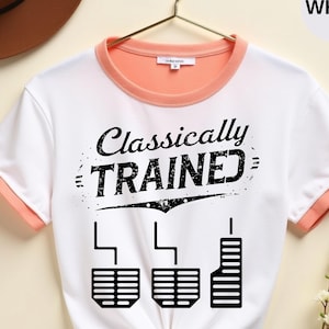 Może przedstawiać: Biała koszulka z brzoskwiniowym wykończeniem i napisem "Classically Trained" nad grafiką pedałów samochodowych. Koszulka wisi na złotym wieszaku. Tekst "WHITE" znajduje się w białym okręgu.