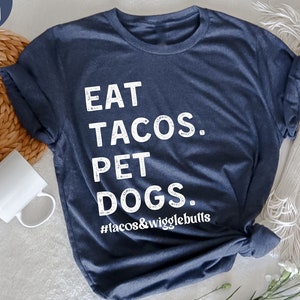 Op de afbeelding: Een heideblauwe t-shirt met de tekst "EAT TACOS. PET DOGS. #tacos&wigglebutts" in wit gedrukt.