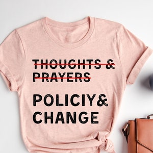 May include: A heather peach t-shirt with the text "THOUGHTS & PRAYERS" crossed out and replaced with "POLICY & CHANGE" in black font. The shirt is displayed on a white surface with accessories.