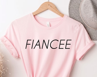 Può includere: Una maglietta rosa chiaro con la scritta "FIANCEE" stampata in nero. La maglietta è appesa a un gancio di legno contro un muro bianco, con un cappello grigio sullo sfondo.