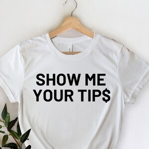 May include: White t-shirt with the text "SHOW ME YOUR TIPS$" in black, hanging on a wooden hanger. A sprig of green leaves and a branch are placed on the lower left side of the shirt.