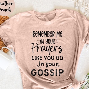 May include: Heather peach-colored t-shirt with the text "Remember Me In Your Prayers Like You Do In Your Gossip" in black. The shirt is folded and tied at the bottom. A white mug and decorative items are in the background.