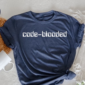 May include: A navy blue t-shirt with the text "code-blooded" in white block letters. The shirt is made of a soft material and is styled with the sleeves rolled up. A white mug and a woven basket are in the background.