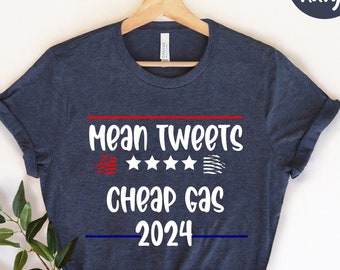 May include: Heather navy t-shirt with the text "Mean Tweets Cheap Gas 2024" in white, red, and blue. The design includes stars and stripes. The shirt is displayed on a wooden hanger.