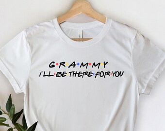 May include: White t-shirt with the text "Grammy I'll Be There For You" in a colorful, stylized font. The shirt is hanging on a wooden hanger, with a sprig of greenery in the lower left corner.