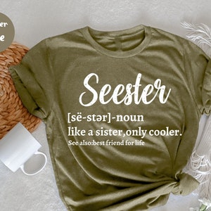 以下が含まれることがあります： 白い筆記体で「Seester」と書かれた緑色のTシャツ。単語の定義は、白い文字で下に書かれています。「[së-stər]-名詞 姉妹のようなもの、ただもっとクール。参照: 一生の親友。」