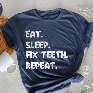 May include: Navy blue heather t-shirt featuring the white text "EAT. SLEEP. FIX TEETH. REPEAT." The shirt is presented on a white surface, accompanied by a white mug, a woven basket, and some greenery.
