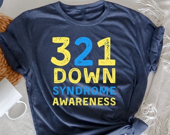 May include: Heather navy t-shirt with the text "321 DOWN SYNDROME AWARENESS" in yellow and blue. The shirt is folded, with a white mug and woven basket in the background. The shirt is made of a soft material.