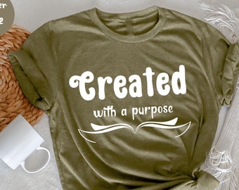 Puede incluir: Camiseta verde oliva con el texto blanco "Created with a purpose". El diseño incluye un gráfico de libro abierto. Una taza blanca y una cesta tejida son visibles en el fondo.