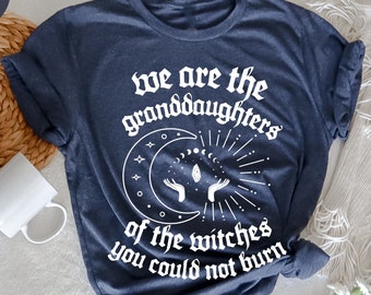 Könnte beinhalten: Heather-Navy-T-Shirt mit weißem Text "we are the granddaughters of the witches you could not burn". Das Design zeigt einen Mond, Sterne und Hände. Eine weiße Tasse und ein geflochtener Korb sind im Hintergrund.