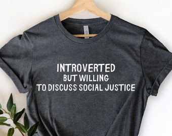 May include: Dark gray t-shirt with the text "INTROVERTED BUT WILLING TO DISCUSS SOCIAL JUSTICE" in white. The shirt is hanging on a wooden hanger against a white background. Greenery is placed on the lower left side of the shirt.