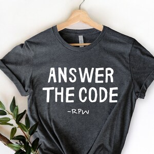 May include: Dark gray t-shirt with the text "ANSWER THE CODE" in bold white letters. Below the text is "-RPW" in smaller white letters. The shirt is hanging on a wooden hanger with a sprig of greenery in the lower left corner.
