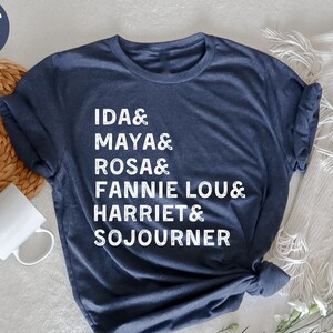 May include: A heather navy blue t-shirt with white text that reads "IDA & MAYA & ROSA & FANNIE LOU & HARRIET & SOJOURNER"