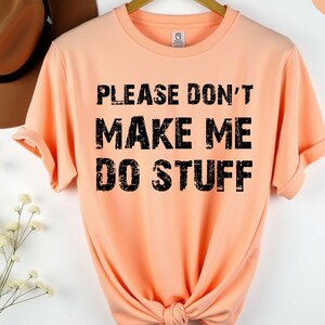 May include: Peach-colored t-shirt with the text "PLEASE DON'T MAKE ME DO STUFF" in black, distressed font. The shirt is knotted at the bottom. A brown hat and flowers are in the background.