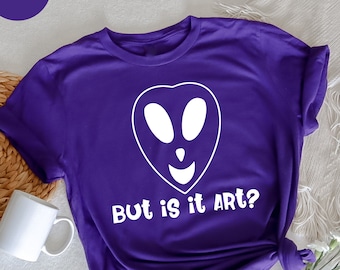May include: A purple t-shirt with a white alien face graphic and the text "But is it ART?" The shirt is a solid purple color and has a crew neck. A white mug and flowers are in the background.