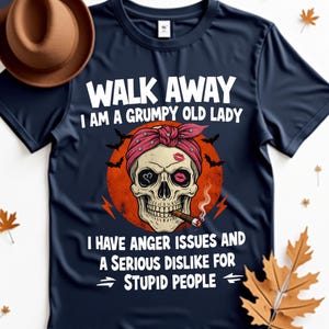Op de afbeelding: Marineblauw t-shirt met de tekst "WALK AWAY I AM A GRUMPY OLD LADY". Het shirt heeft een schedelafbeelding met een rode bandana, een kusafdruk en een brandende sigaar. Het shirt heeft ook de tekst "I HAVE ANGER ISSUES AND A SERIOUS DISLIKE FOR STUPID PEOPLE".