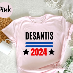 以下が含まれることがあります： 黒色の「DESANTIS」という文字、2本の青い水平線、赤色の「2024」の数字、2つの黒い星がプリントされた、明るいピンク色のTシャツ。「Pink」という文字が左上のピンク色の円の中に入っています。