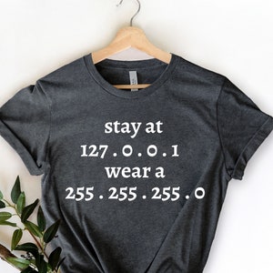 Könnte beinhalten: Dunkelgraues T-Shirt mit dem weißen Text "stay at 127.0.0.1 wear a 255.255.255.0". Das Shirt hängt an einem Holzbügel. Grüne Blätter befinden sich in der linken unteren Ecke.
