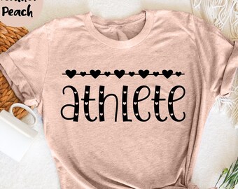 May include: Heather peach-colored t-shirt with the word "athlete" in black script. The word is decorated with small black hearts. Above the word is a line of black hearts. The shirt is styled with rolled-up sleeves.