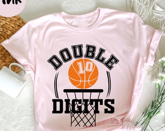 Könnte beinhalten: Hellrosa T-Shirt mit dem schwarzen Schriftzug "DOUBLE DIGITS". Ein orangefarbener Basketball mit der Nummer 10 befindet sich in der Mitte. Das Shirt hat eine Basketballkorb-Grafik und ist unten geknotet.
