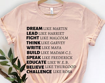 Könnte beinhalten: Ein hellrosa T-Shirt mit schwarzem Text. Der Text enthält die Wörter "DREAM, LEAD, FIGHT, THINK, WRITE, BUILD, SPEAK, EDUCATE, BELIEVE, CHALLENGE" gefolgt von Namen wie Martin, Harriet, Malcolm und anderen. Das T-Shirt hängt an einem Holzbügel.