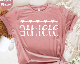 May include: Heather mauve t-shirt with the word "athlete" in white script, decorated with a row of hearts. The shirt is folded and tied at the bottom. A white mug and flowers are in the background.