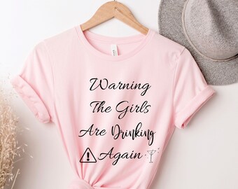 Pode incluir: T-shirt rosa claro com o texto preto "Warning The Girls Are Drinking Again". A camisa está pendurada em um cabide de madeira. Um chapéu cinza é visível no fundo.