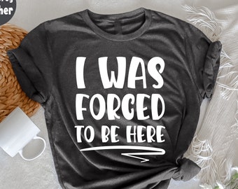 Puede incluir: Camiseta gris oscuro jaspeado con el texto blanco "I WAS FORCED TO BE HERE". Las mangas están dobladas y la cintura anudada. Una taza blanca y una cesta tejida están en el fondo.